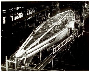 Overhead shot of hull being planked inside the boatshop, men at work rivetting the steel plates onto the steel frames.